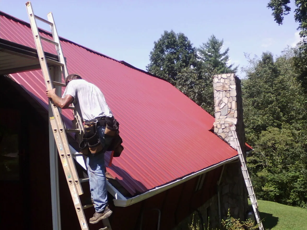 Roofer on a ladder beside a red metal roof and stone chimney — Soffit & Fascia Repair by Roofing Brothers in Hibernia